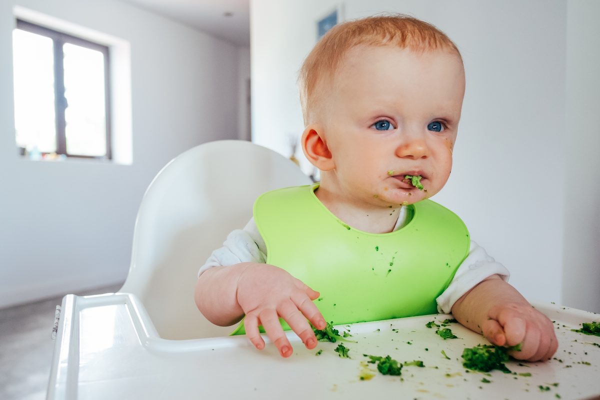 Baby in a high chair feeding themselves cooked vegetables