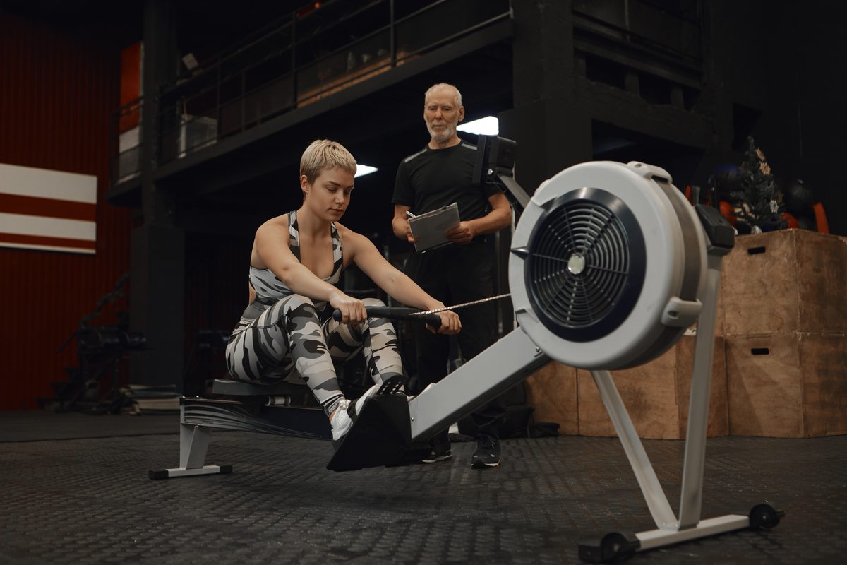 Coach sportif avec un bloc-notes observant une cliente sur un rameur