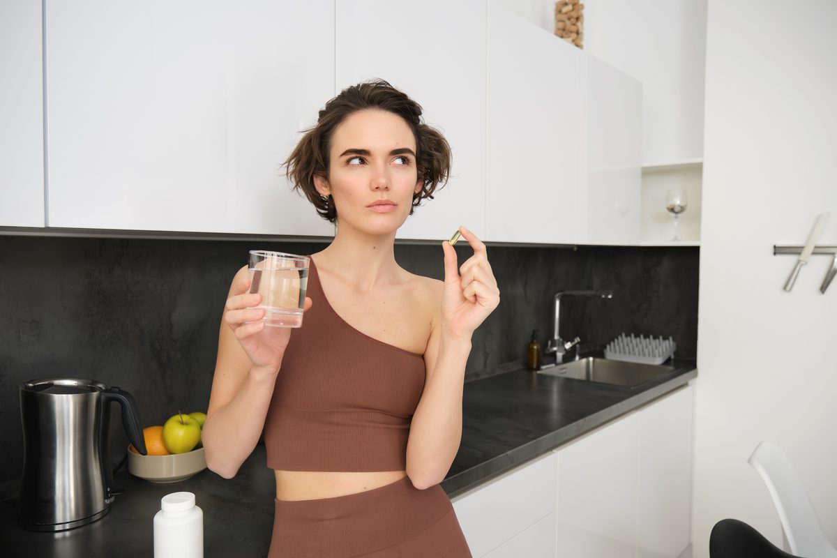 Femme en tenue de sport tenant une gélule et un verre dans sa cuisine
