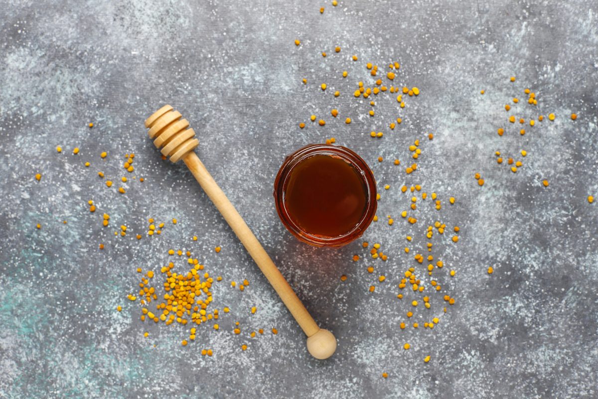 Small pot of honey and wooden spoon surrounded by grains of pollen