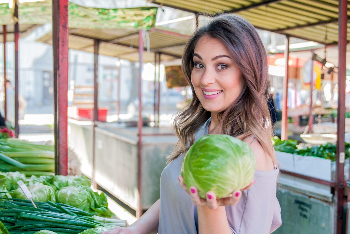 Young woman holding a green cabbage in front of a vegetable stall at the market